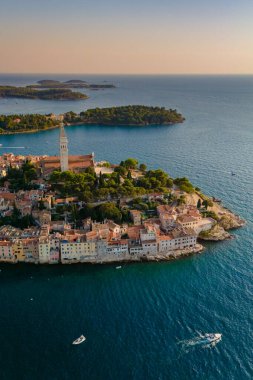 Güneş batarken Rovinj 'in sakin suları parıldıyor ve tarihi binalar ve yemyeşil alanlar üzerine sıcak bir parıltı saçıyor. Tekneler yavaşça süzülüyor, sakin sahil atmosferini güçlendiriyor..