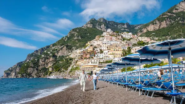 İki kişi, Amalfi sahilinin çakıllı sahillerinde el ele yürürken, Positano 'nun canlı uçurum kenarındaki evleri açık mavi gökyüzünün altında görkemli bir şekilde yükseliyor..