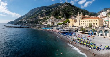 Güneş banyosu, Amalfi Sahili 'nin canlı kıyılarında, kumsalı süsleyen renkli şemsiyelerle yapılmaktadır. Göz kamaştırıcı uçurumlar parlak mavi gökyüzünün altında muhteşem bir şekilde arka planda yükseliyor. Amalfi İtalya