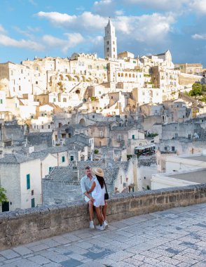 Bir çift, gün batımının sıcacık parıltısında güneşlenen taş bir duvarda oturuyor. Aşağıda, Matera İtalya 'nın tarihi şehir manzarası, antik mimarisini ve dolambaçlı yollarını gözler önüne seriyor..