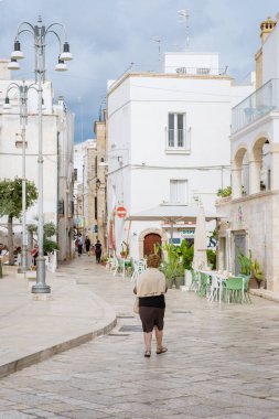 Ostuni İtalya 18 Eylül 2024, Puglia 'nın sakin sokaklarında gezen yalnız bir şahıs, tuhaf binalar ve davetkar kafelerle çevrili. Yumuşak bulutlar üzerimizden geçerken atmosfer sakin.