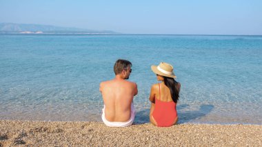 Two individuals enjoy a peaceful moment together, seated on a pebbly beach while soaking in the stunning views of clear turquoise waters under a bright summer sky. Zlatni Rat Beach clipart