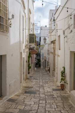 İtalya 'nın Puglia şehrinde 18 Eylül 2024' te sakin, dar bir caddede büyüleyici beyazlatılmış binalar ve davetkar dükkanlar sergileniyor. Işıkların dizeleri yukarıda asılı,