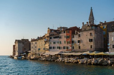 Rovinj 'in canlı binaları kıyı şeridine yapışıyordu. Renkli cepheleri sıcak akşam ışığında parlıyordu. Dalgalar yavaşça kıyıya vururken yerel halk ve ziyaretçiler sakin atmosferin tadını çıkarıyorlar..