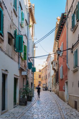 Yalnız bir figür, Rovinj 'de güzel bir kaldırım taşı sokakta geziniyor. Binaların canlı renkleri ve yemyeşil yeşil berrak mavi gökyüzünün altında büyüleyici bir atmosfer yaratır..