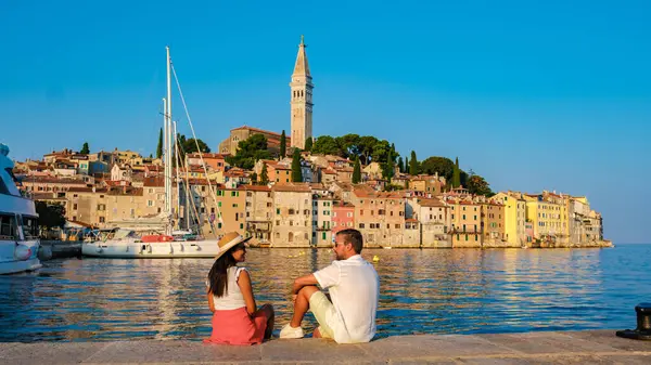 Güneş ufkun altına batarken, Rovinj 'deki rıhtımda büyüleyici ve renkli mimari ile çevrili bir çift rahatlıyor. Sakin sular günbatımının canlı renklerini yansıtır..