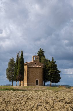 Dramatik bir gökyüzü altında, Kıbrıs ağaçlarının arasında altın tarlalarla çevrili, sakin bir sonbahar öğleden sonrasında Toskana 'nın ebedi güzelliğini yakalayan tuhaf bir şapel duruyor..