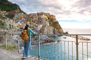 Bir gezgin, sakin denizin yanındaki kayalıklara tünemiş renkli binalarıyla büyüleyici Manarola 'ya bakar. Sakin dalgalar alacakaranlığın yumuşak ışıltısının altında parıldarken..