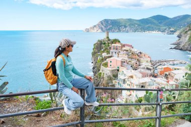 Bir gezgin tırabzanda oturup Cinque Terre 'nin çarpıcı sahil şeridine bakar. Uçurumlara tünemiş canlı evler ve ışıl ışıl deniz manzaralı bir zemin oluşturuyor, Vernazza İtalya