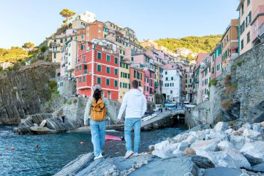 Bir çift, yamaçtan aşağı çağlayan canlı binalarla çevrili kayalık sahilde el ele duruyor. İtalya Cinque Terre 'de Riomaggiore