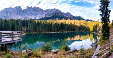 Early morning light casts a serene glow on the emerald waters of a mountain lake, surrounded by towering pine trees and dramatic mountain peaks of Lake Carezza or Karersee Dolomites in Italy. clipart