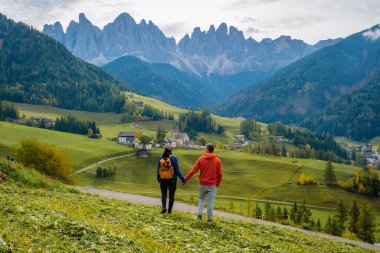 Dolomitlerin göz kamaştırıcı sonbahar manzaralarıyla çevrili dolambaçlı bir yolda el ele yürüyen bir çift. Santa Maddalena Val di Funes İtalya 'daki büyüleyici köylerin arkasında görkemli dağlar yükseliyor.