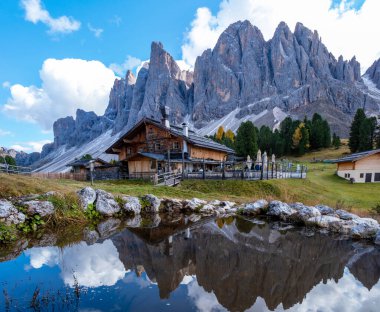 Çarpıcı Dolomitler, arka plandaki heybetli dağ sıralarını yansıtan sakin bir göletin yanında duran tuhaf ahşap bir kulübe. İtalya 'da Geisleralm Dolomitleri Val Di Funes Adolf Munkel Patikası