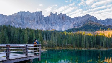 A solitary traveler stands on a wooden bridge, gazing at the serene turquoise waters reflecting the majestic Dolomites. Lake Carezza or Karersee Dolomites in Italy. clipart