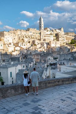Bir çift, Matera İtalya 'nın nefes kesen manzarasından büyülenmiş taş bir duvarın yanında duruyor. Eski taş binaları ve yüksek binaları öğleden sonra güneş ışığında yıkanmış..