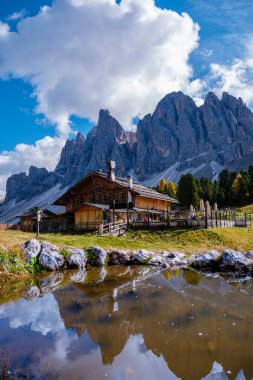 Nestled against the stunning backdrop of the Dolomites, a charming wooden cabin stands proudly as autumn colors surround it. Geisleralm Dolomites Val Di Funes in Italy A clipart