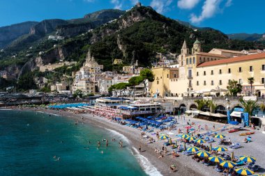 Güneş Amalfi Sahili 'ni aydınlatırken turistler çakıllı bir sahilde renkli şemsiyelerin altında güneşleniyor. Manzara kayalıkları ve büyüleyici binalar bu Akdeniz kaçışı için çarpıcı bir zemin oluşturuyor..