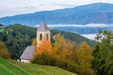 Resmedilmeye değer Dolomitler 'in arasında, canlı sonbahar yapraklarının arasında, şafak vakti sislerin yavaşça vadiden yükselmesiyle dingin ve büyüleyici bir atmosfer yaratan tarihi bir kilise bulunur..