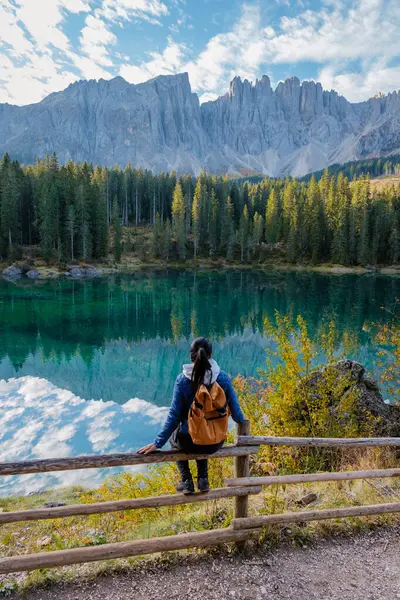 Yanlız bir yürüyüşçü ahşap bir çitin yanında duruyor, İtalya 'daki bereketli ormanlar, Carezza Gölü veya Karersee Dolomitleri ile çevrili berrak bir göldeki engebeli dağların nefes kesici yansımasına bakıyor..