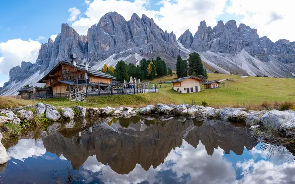 Dolomitlerin nefes kesen zirveleri mavi bir gökyüzüne karşı görkemli bir şekilde yükseliyor, dingin bir gölette güzel bir şekilde yansıyor. Yeşilliğe gömülü ahşap kulübeler, İtalya 'da Geisleralm Dolomitleri Val Di Funes