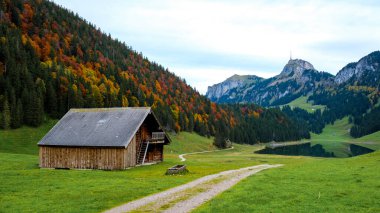 Nestled amidst vibrant autumn colors, a charming wooden cabin stands peacefully by Falensee, surrounded by majestic mountains and lush green meadows under an expansive sky.