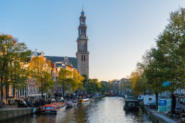 Amsterdam 23 Ekim 2024, altın güneş ışığı kanalları yıkarken, canlı sonbahar yaprakları Amsterdam 'ın ikonik kulesini çerçeveliyor. İnsanlar suyun üzerinde yürüyor, şehrin huzurlu güzelliğinin tadını çıkarıyorlar..
