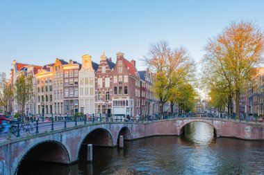 Dondurucu sonbahar esintisinde altın yapraklar dalgalanıyor bisikletçiler Amsterdam 'daki huzurlu köprüleri geçerken, büyüleyici kanal evleriyle çevrili, mevsim güzelliğini ve dinginliğini yansıtıyor..