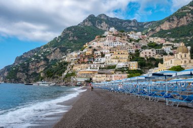 Visitors wander the beautiful pebble beach of the Amalfi Coast, surrounded by charming hillside homes and stunning views. The azure waters gently lap against the shore under a partly cloudy sky. clipart