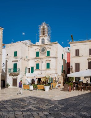 Polignano a Mare Italy 18 Eylül 2024, Puglia 'da güneşli bir gün ziyaretçilerin yeşil panjurlarla süslenmiş, sıcak atmosferin keyfini çıkardıkları ilginç binaların önünden geçtikleri canlı bir meydanı ortaya çıkardı.