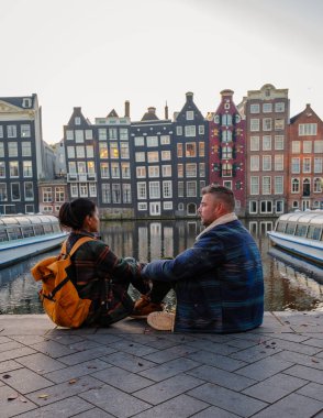 Hollanda, Amsterdam 'daki renkli tarihi binalarla dolu sakin kanallardan yansıyan altın sonbahar ışıklarının tadını çıkaran iki kişi kaldırım taşı yolunda birlikte oturuyorlar..