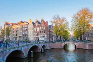 Dondurucu sonbahar esintisinde altın yapraklar dalgalanıyor bisikletçiler Amsterdam 'daki huzurlu köprüleri geçerken, büyüleyici kanal evleriyle çevrili, mevsim güzelliğini ve dinginliğini yansıtıyor..