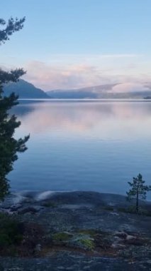 Sakin bir göl kenarında dingin bir sabah belirir, yumuşak dalgalar şafağın canlı renklerini yansıtır. Norveç 'teki yumuşak bulutlardan oluşan bir battaniyenin altında görkemli dağlar beliriyor.