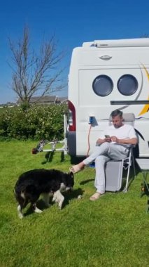 Bir insan açık havada sakin bir öğleden sonranın keyfini çıkarırken, canlı bir köpek açık mavi bir gökyüzünün altında hevesle etkileşime geçerken, bir karavan yanında rahat bir şekilde oturuyor. Texel Hollanda