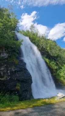 Çarpıcı bir şelale engebeli kayalıklardan süzülüyor yemyeşil sularla çevrili. Norveç 'te doğa harikalar yaratırken, parlak mavi gökyüzü sakin atmosfere katılıyor..