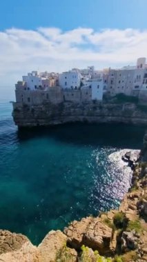 Polignano a Mare Puglia Italya 'nın resimsel güzelliğini keşfedin. Dalgalar kayalık oluşumlara hafifçe çarparak açık mavi gökyüzünün altındaki kayalıklara tünemiş büyüleyici, renkli binaları ortaya çıkarıyor..