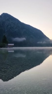 Sabahın erken saatlerinde Plansee Avusturya Gölü 'nün üzerinden yükselen ve görkemli dağları ve ağaçların arasına sıkışmış huzurlu bir kulübeyi yansıtan sisi hissedin.. 