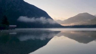 Sabahın erken saatlerinde durgun gölün üzerinde sakin bir parıltı oluşur. Bu, yüksek dağları mükemmel bir şekilde yansıtır. Sis yavaşça suyun üzerinde süzülüyor, Plansee Austria