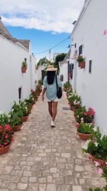 Bir gezgin, canlı saksılarla çevrili, güzel bir kaldırım taşı yolu boyunca dolanıyor. Beyazlatılmış duvarlar gök kubbenin altında dingin bir atmosfer yaratıyor, Alberobello Puglia İtalya