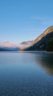 Sakin bir ortamda, şafak dingin bir göl üzerinde doğuyor, etrafındaki dağları yansıtıyor ve yemyeşil. Sakin sular erken ışığı yansıtıyor ve barışçıl bir atmosfer yaratıyor. Plansee Avusturya