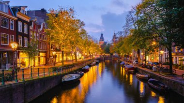 Alacakaranlık Amsterdam 'ın üzerine çöktüğünde, su yolları rahat ışıkların yansımalarıyla parıldıyor. Ağaçlar sonbahar renkleriyle dolup taşar, büyüleyici kanallar boyunca pitoresk bir ortam yaratır..
