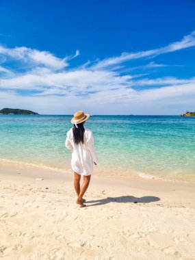 A serene figure stands barefoot on the soft sand of Samae San Island, gazing at the sparkling turquoise sea. The sun illuminates a clear blue sky, creating a perfect tropical escape. clipart