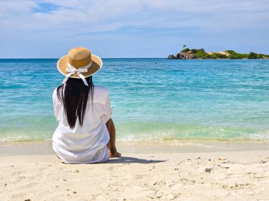 A peaceful moment at Samae San Island, Thailand, captures a woman in a sunhat savoring the stunning ocean view. Gentle waves lap at the shore, creating a tranquil atmosphere on this idyllic beach day. clipart