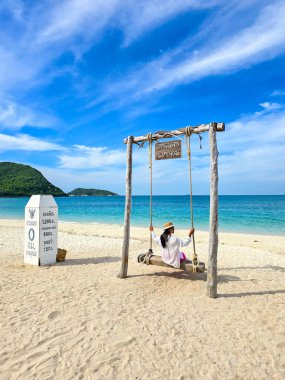 Samae San Island, a woman visitor swings gently over golden sands, with turquoise waters and lush hills in the background under a bright blue sky. ( Translation of Thai text: Samae San Island ) clipart