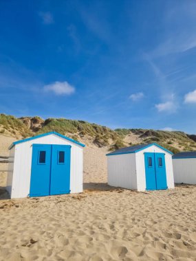 Mavi kapıları olan iki büyüleyici sahil kulübesi yumuşak kumda yuvalanmış, nazik kum tepeleriyle çevrili. Yukarıdaki parlak mavi gökyüzü sakin kıyı atmosferini yükseltiyor, Teksas Hollanda
