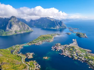 Vibrant blue waters reflect the sky as a picturesque village nestles between towering mountains and scenic coastlines. Lush greenery covers the landscape, enhancing the natural beauty. Lofoten Norway clipart