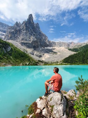 Bir adam, Dolomitlerdeki görkemli dağlarla çevrili nefes kesici bir turkuaz gölün kenarındaki kayanın üzerinde oturuyor ve doğanın dingin güzelliğinin tadını çıkarıyor, lago Di Sorapis Dolomites İtalya