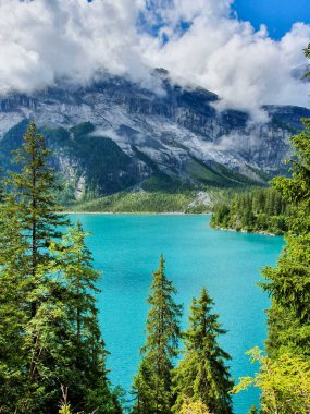 Çarpıcı bir turkuaz göl mavi gökyüzünü ve uzun yeşil ağaçlarla çevrili kabarık bulutları yansıtır. Arka planda heybetli dağlar. Oeschinensee İsviçre