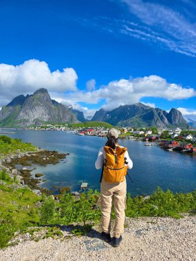 Bir yürüyüşçü, Lofoten Norveç 'teki gür bir arazinin kenarında huzur içinde durur. Büyüleyici dağlara ve aşağıdaki tuhaf köye hayret eder.. 