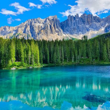 Sakin bir alp gölü, yemyeşil yemyeşil ağaçlarla çevrili çarpıcı turkuaz suları sergiliyor, bu da güneş ışığıyla yıkanan yüksek dağları yansıtıyor. Karersee veya Lago Di Carezza Dolomites İtalya