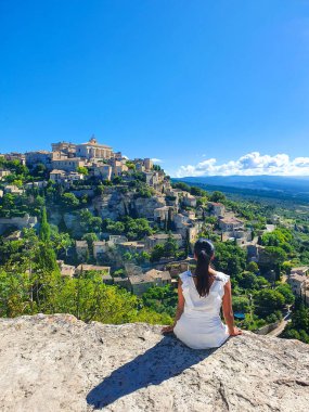 A young woman in a white dress sits peacefully on a rocky ledge, gazing at the picturesque village of Gordes. The bright sky enhances the lush greenery, Gordes Luberon Provence clipart